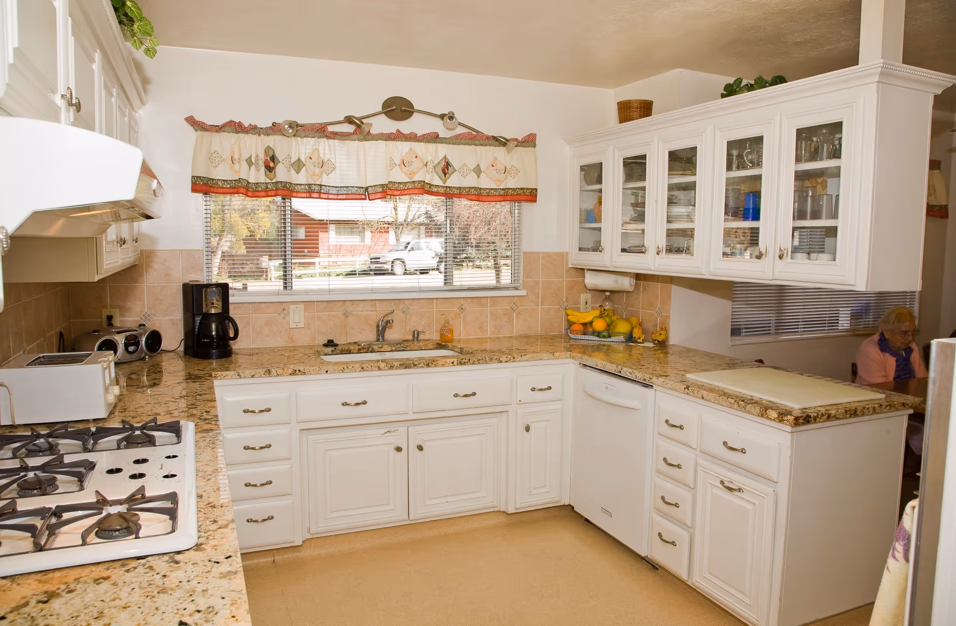 A bright kitchen with white cabinets and granite countertops. There is a gas stove on the left, a coffee maker, toaster, and a fruit basket on the counter. A window with a decorative valance overlooks the outside. In the background, an elderly person is seated at a table partially visible through an open doorway.