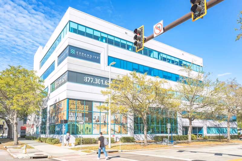 Modern glass-front building with trees, pedestrians, and blue sky along a city street corner.