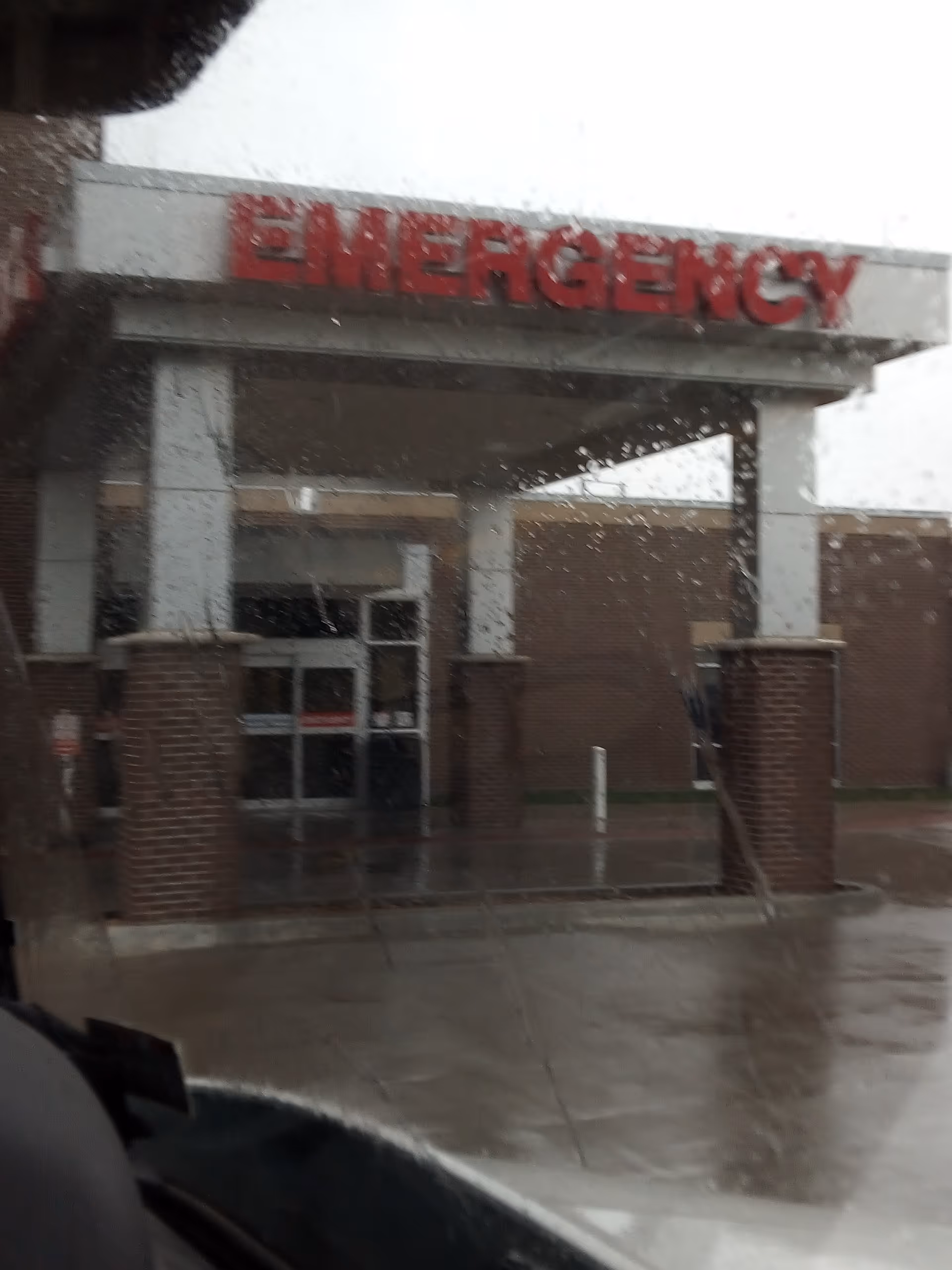 Front view of a hospital entrance with a red EMERGENCY sign and wet pavement.