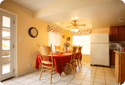 Bright dining room with a red tablecloth, wooden chairs, and a kitchen area.