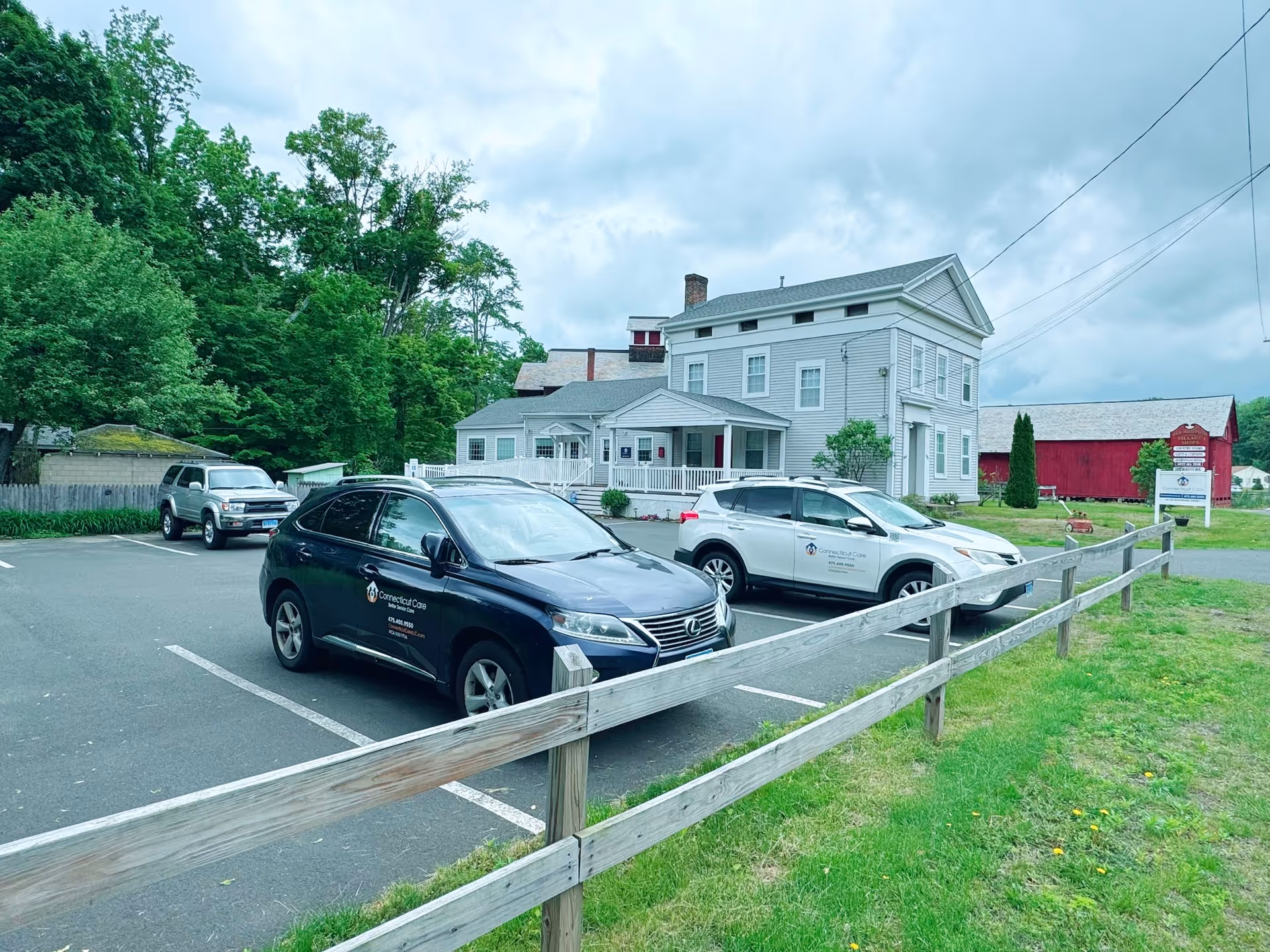Exterior view of Connecticut Care facility with parked cars, wooden fence, and surrounding trees.