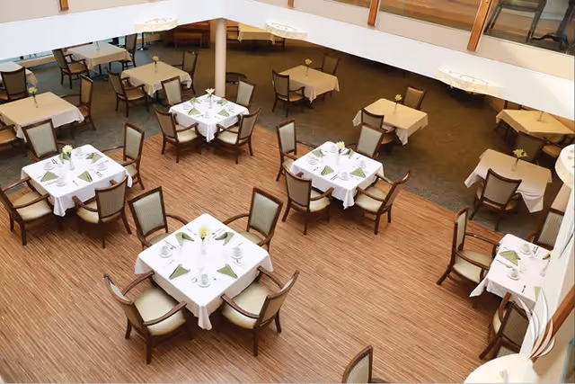 A spacious dining room with multiple square tables covered with white tablecloths, each set with plates, cups, and green napkins. The chairs around the tables have wooden frames and cushioned seats. The floor is a combination of wood and carpet, and the room is well-lit with natural light from large windows.