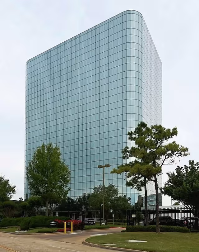 Tall curved glass office building with trees and a parking lot in front.
