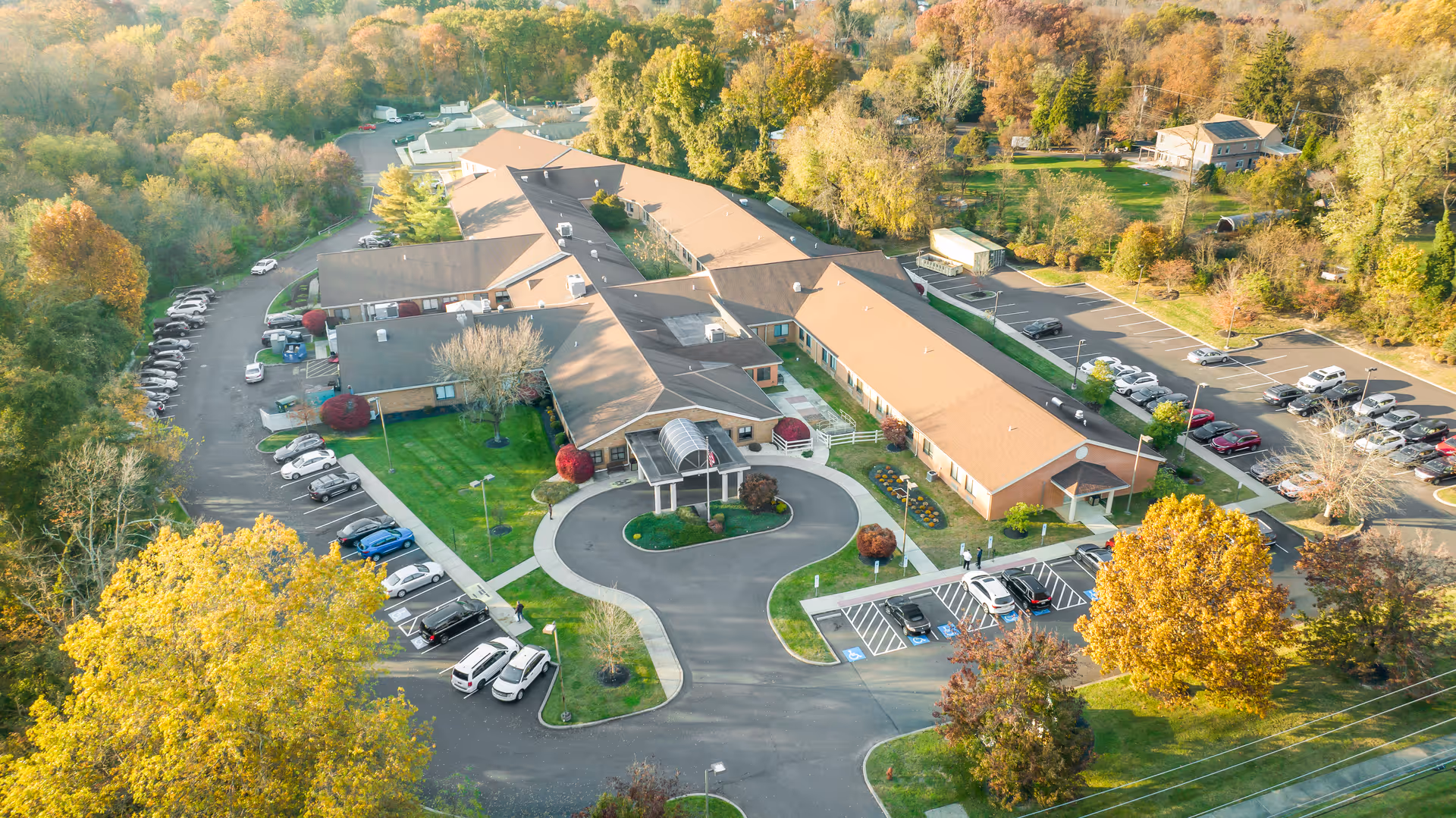 Aerial view of Yardley Rehabilitation and Healthcare Center surrounded by trees with autumn foliage. The building has a tan roof and is shaped in a U-formation with a circular driveway and parking lots on both sides. Several cars are parked in the lots and the surrounding area is lush with greenery.