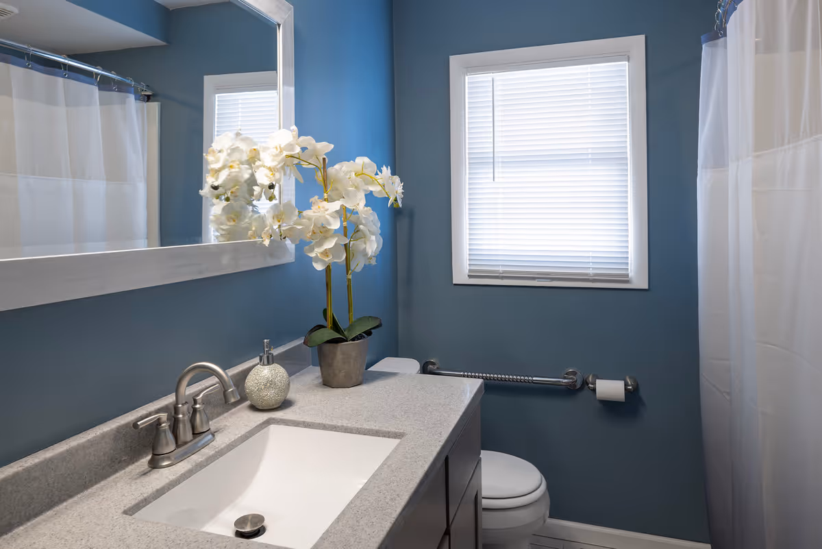 Blue bathroom with sink, mirror, window blinds, orchid plant, toilet, grab bar, and shower curtain.