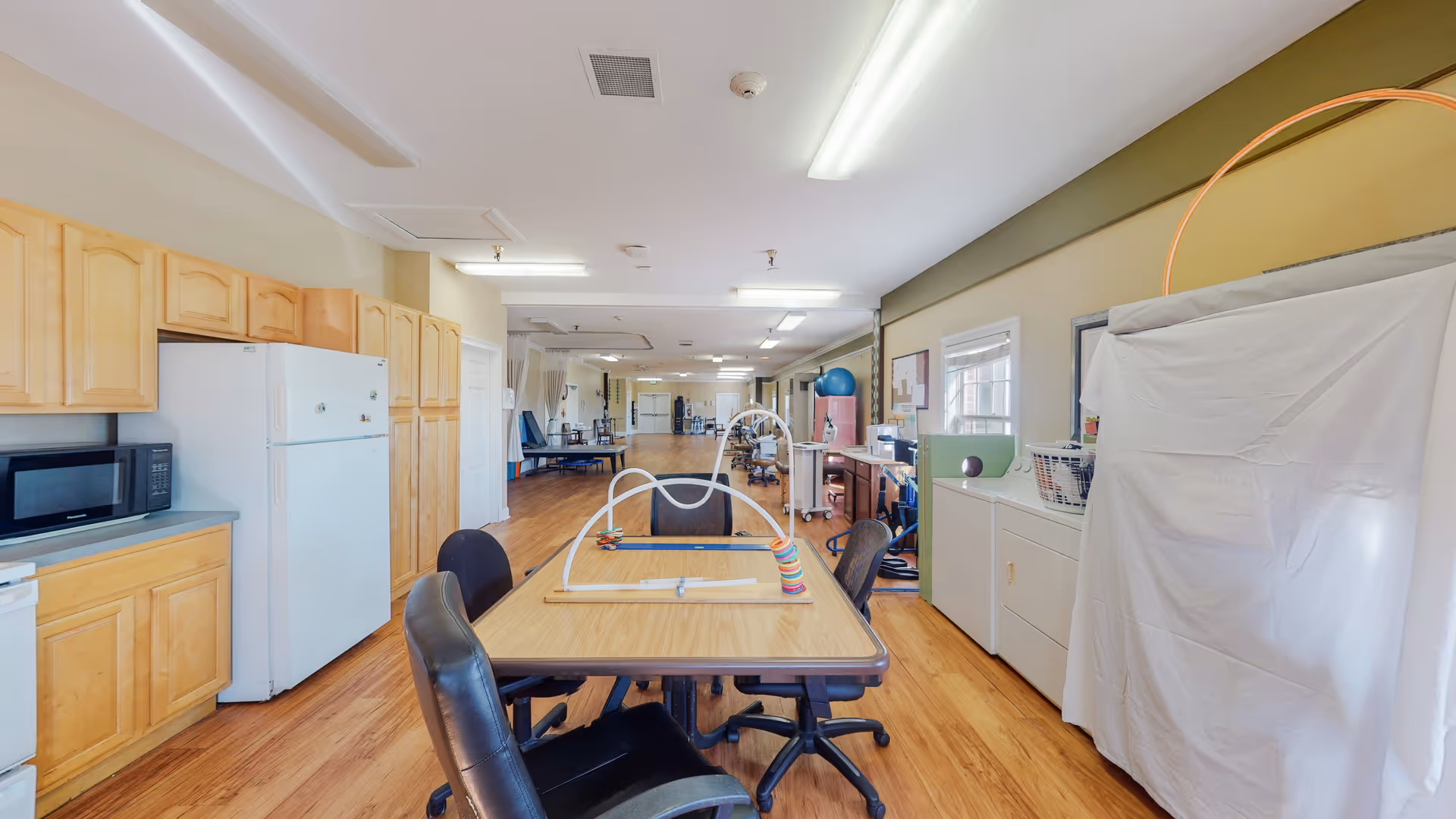 Long communal activity room with a central table, kitchenette, and covered partition.