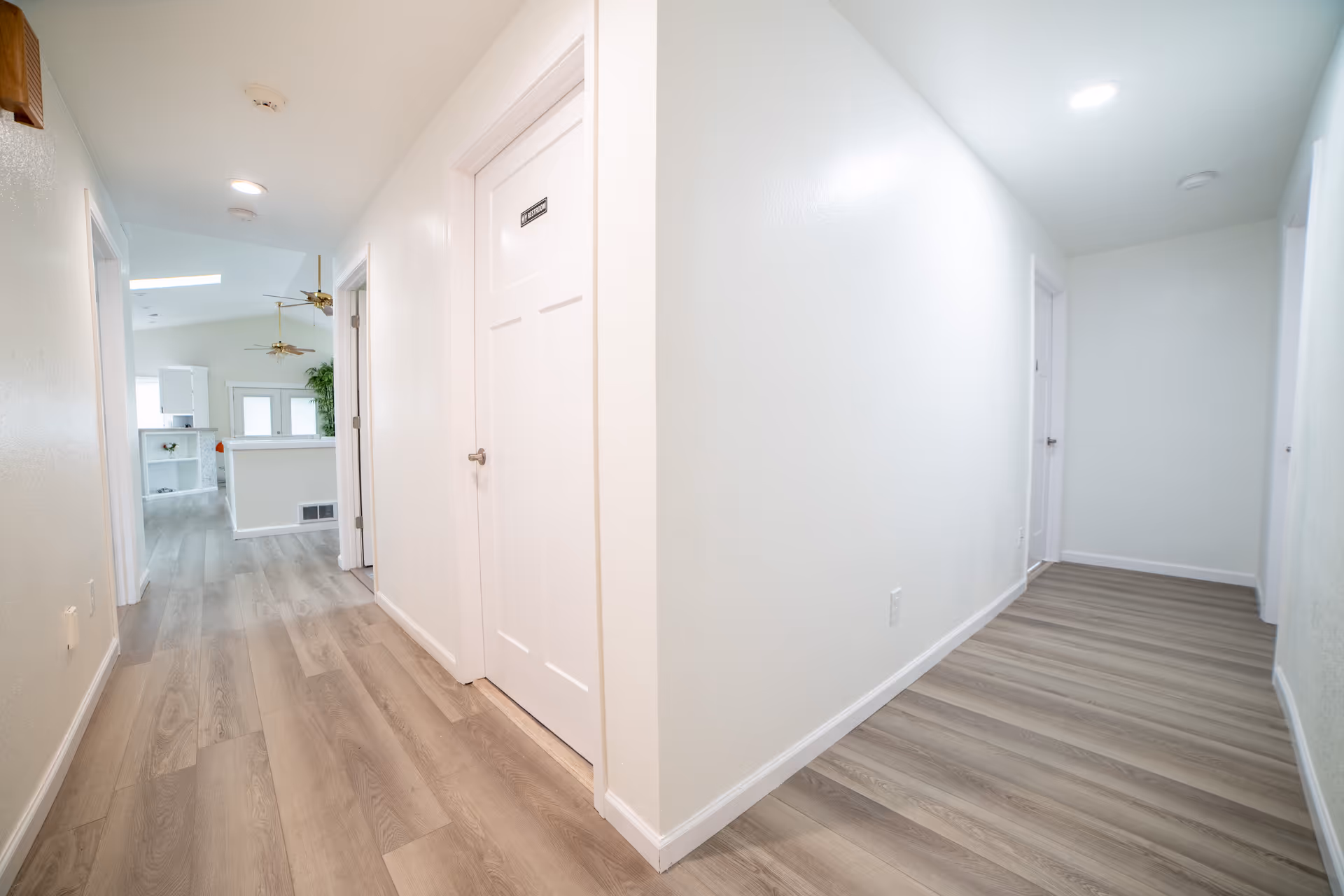 Bright hallway with white walls, doors, and light wood flooring in a senior living home.