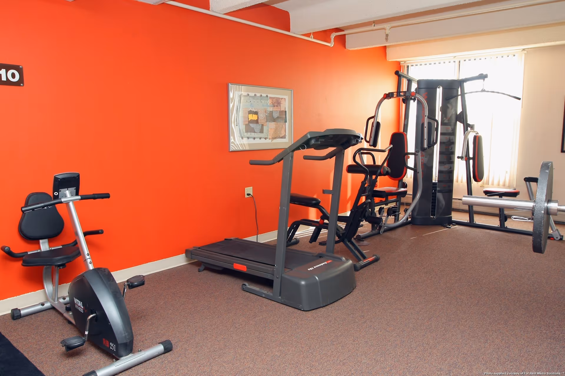 Indoor fitness room with exercise equipment including a stationary bike, treadmill, and multi-function weight machine against an orange wall with a framed abstract artwork.