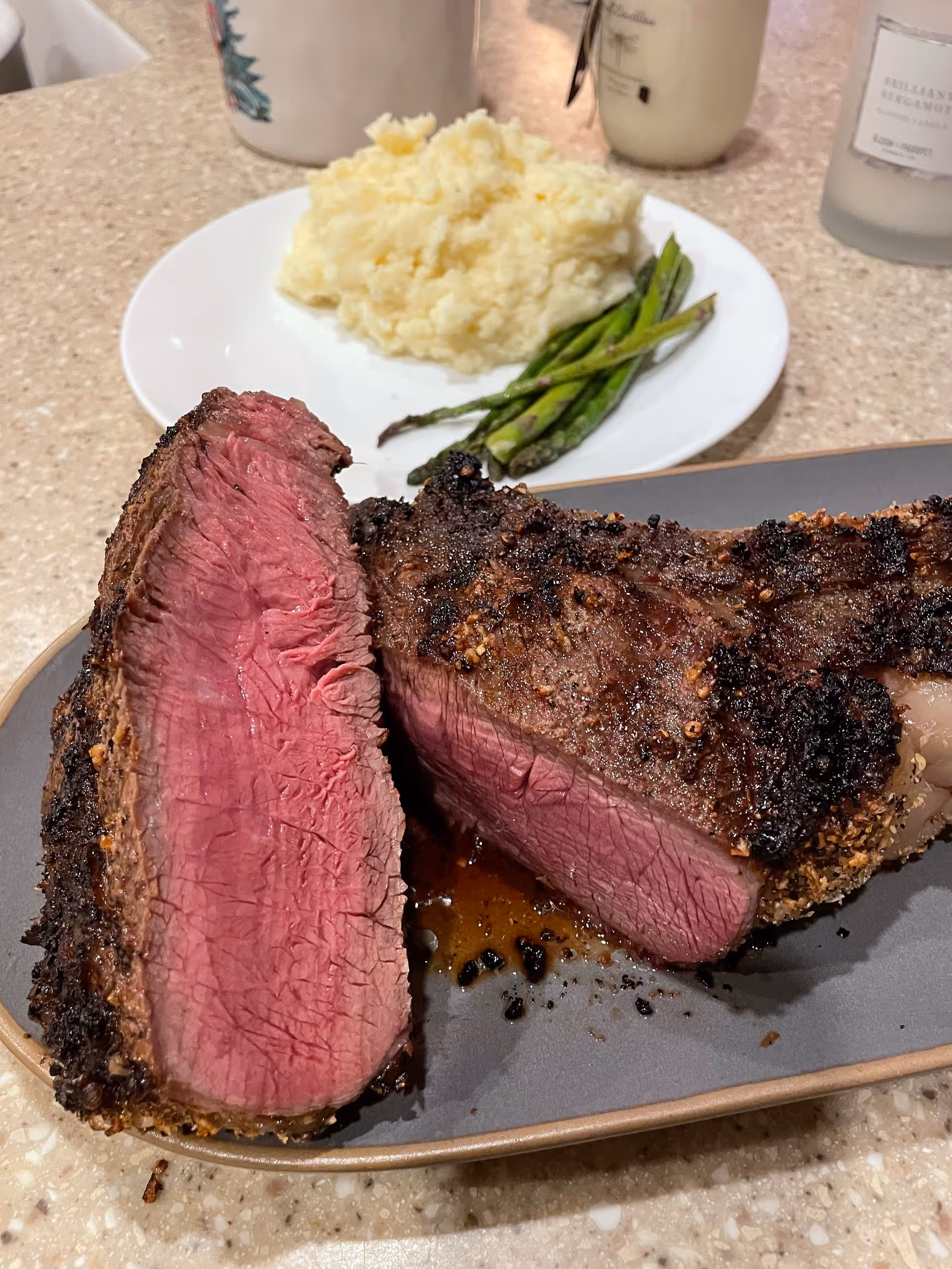 A close-up view of a sliced medium-rare steak on a gray plate with a white plate in the background holding mashed potatoes and asparagus on a countertop.