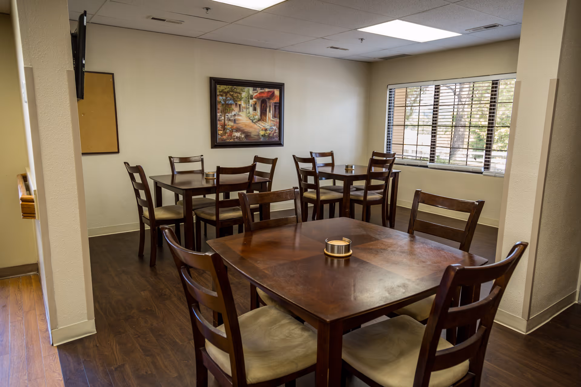 Interior dining area with wooden tables and chairs, window blinds, and framed wall art.