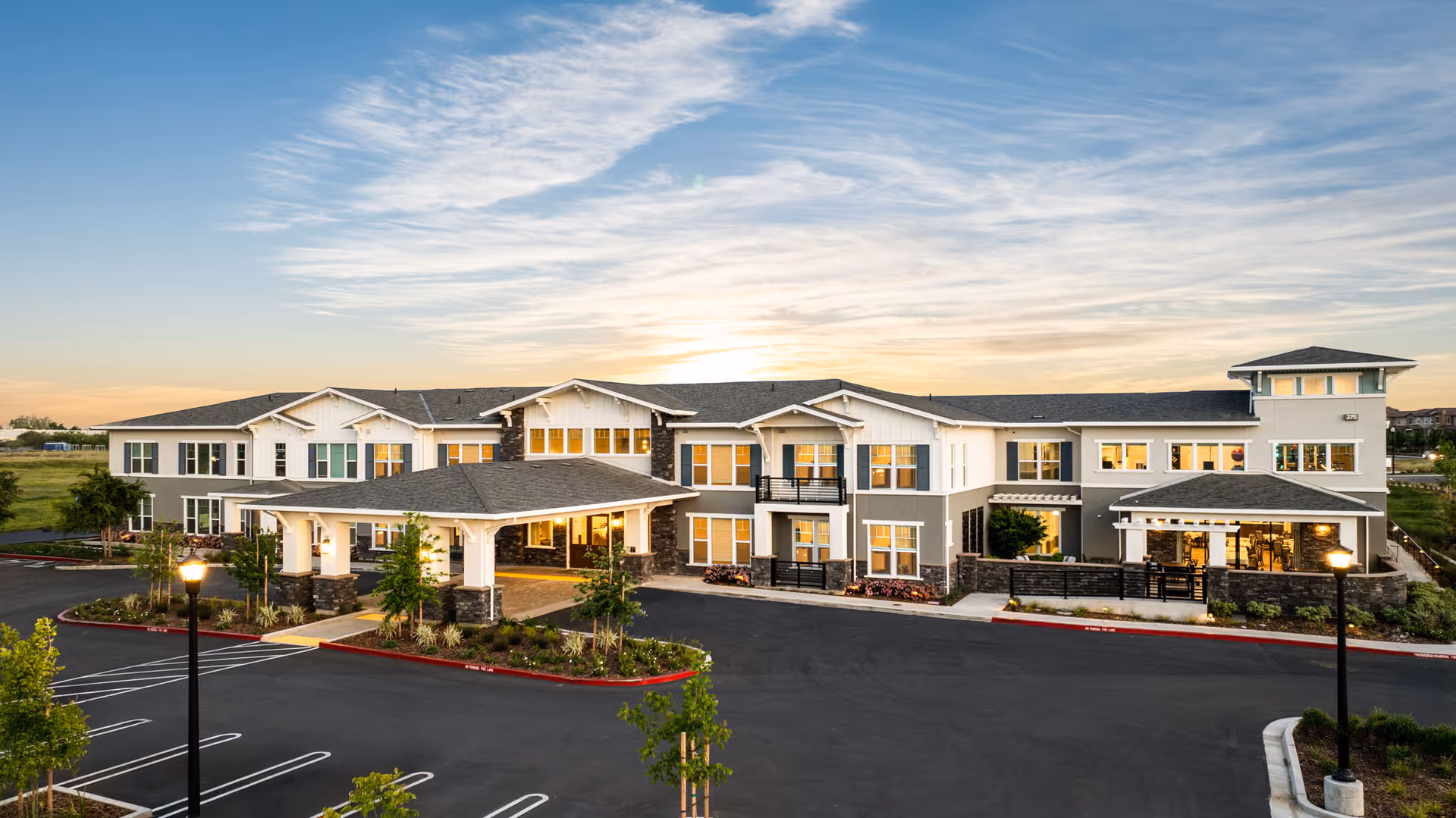 Exterior of a modern two-story senior living facility at sunset with a large parking lot.