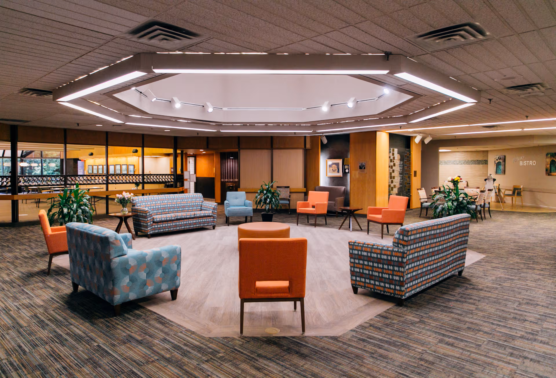 Spacious senior living lounge with colorful seating arranged around a central octagonal lighting fixture.