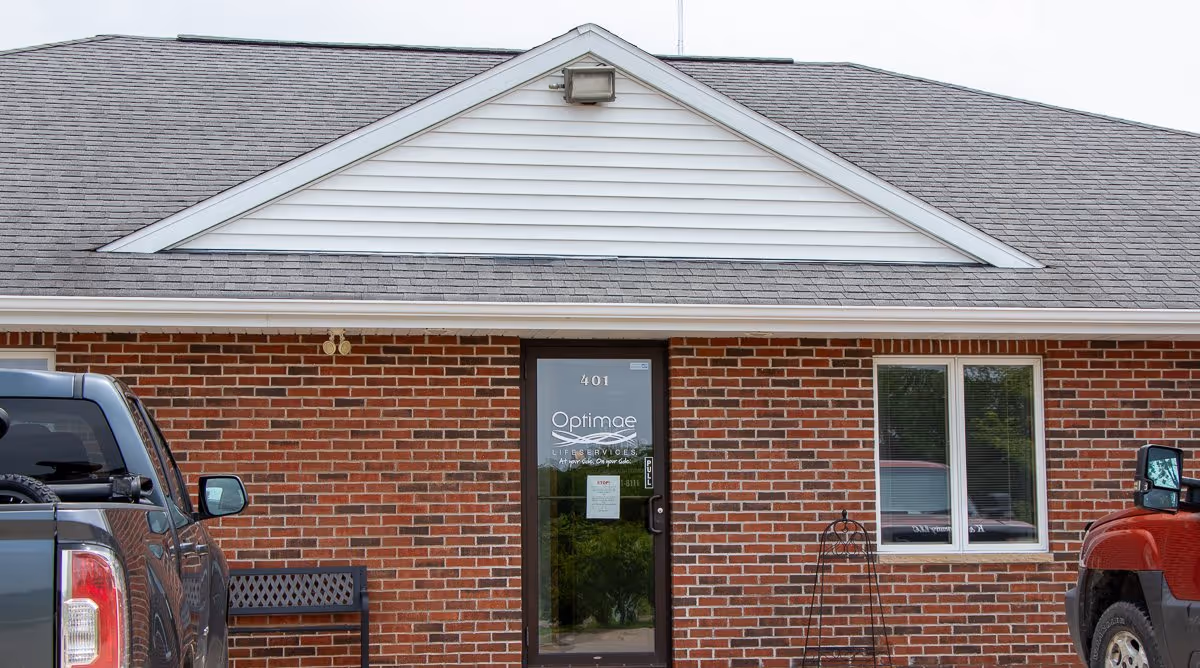 Brick building facade with an entrance labeled Optimae, flanked by vehicles and a window.