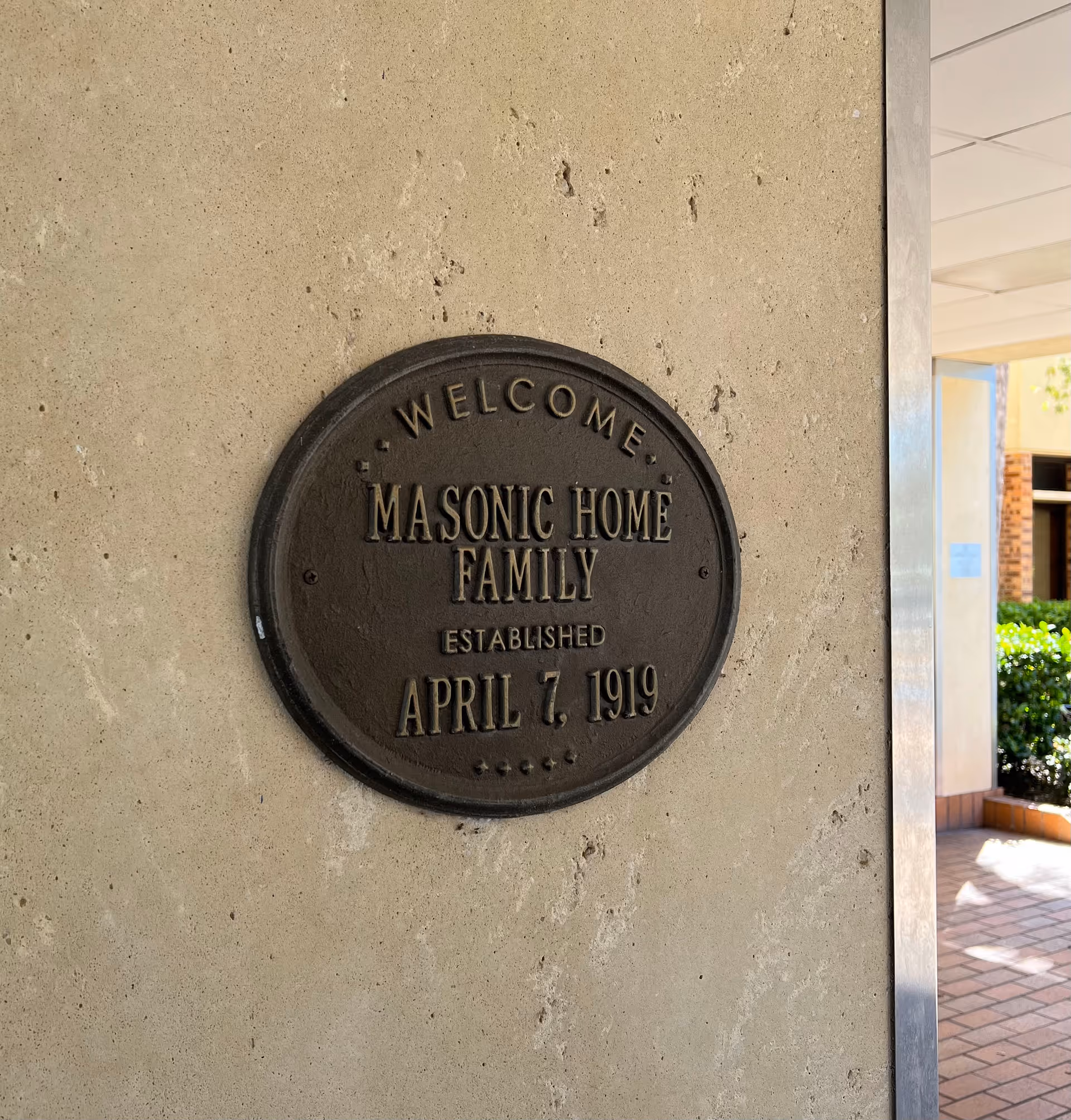 Metal round plaque reading Welcome Masonic Home Family on beige exterior wall.