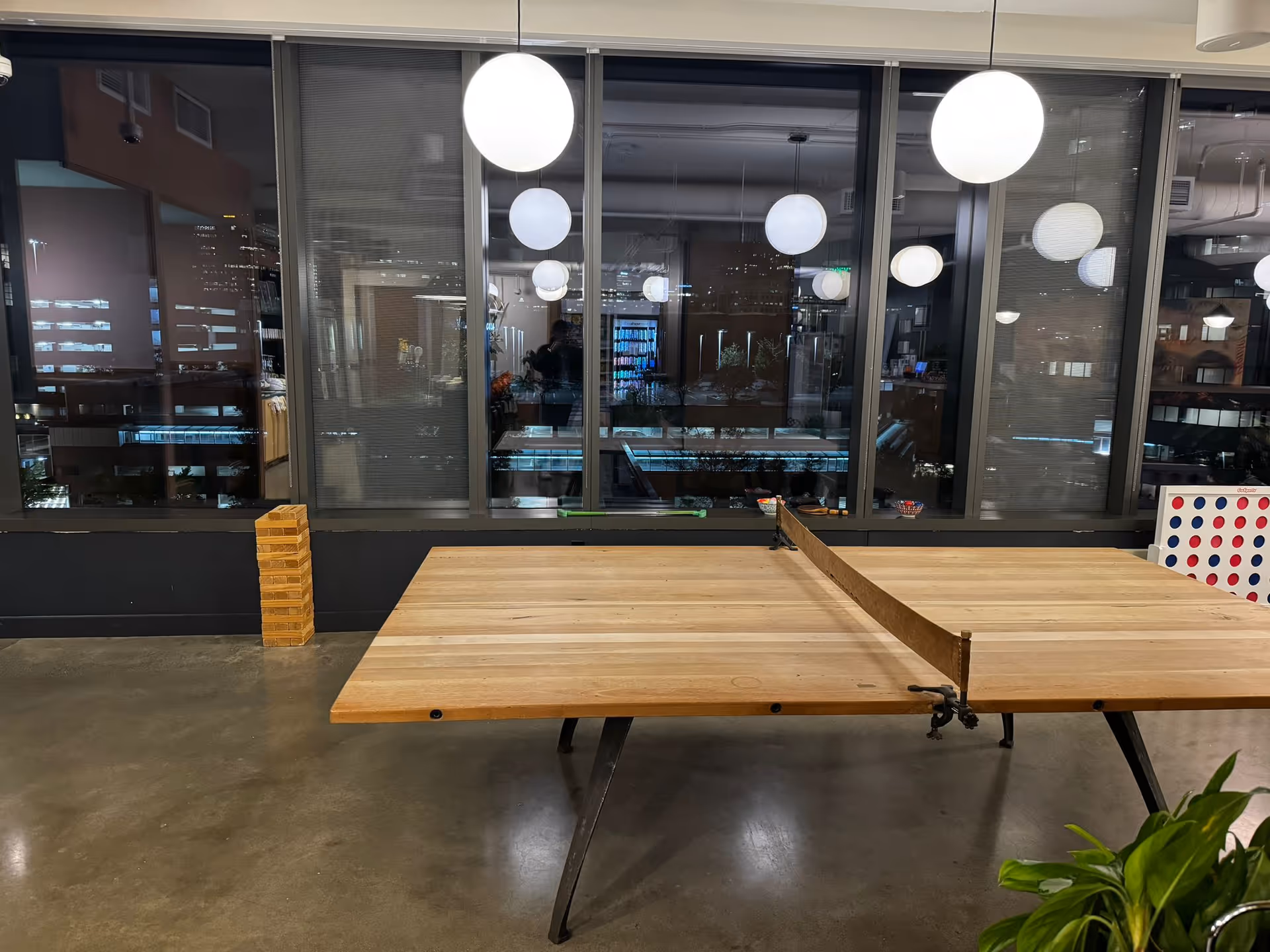 Empty modern lounge with a large wooden table, pendant lights, and city skyline visible through windows.