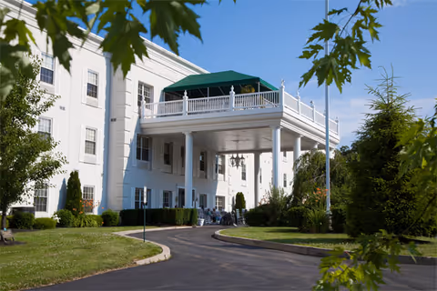 White multi-story senior living building with a covered porte-cochère entrance, upper balcony with a green awning, driveway, and surrounding landscaping.