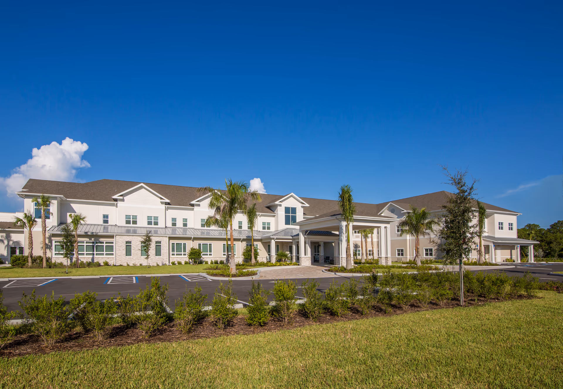 Two-story light-colored senior living building with a covered entrance, palm trees, landscaped grounds, and a parking lot under a clear blue sky.