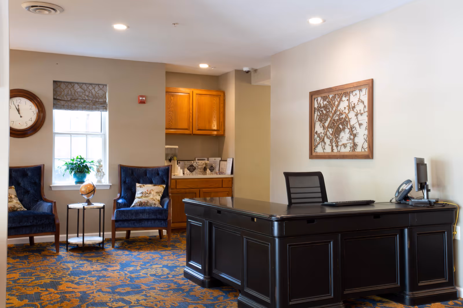 A spacious lobby area with a dark reception desk, blue chairs, and kitchenette in the background.