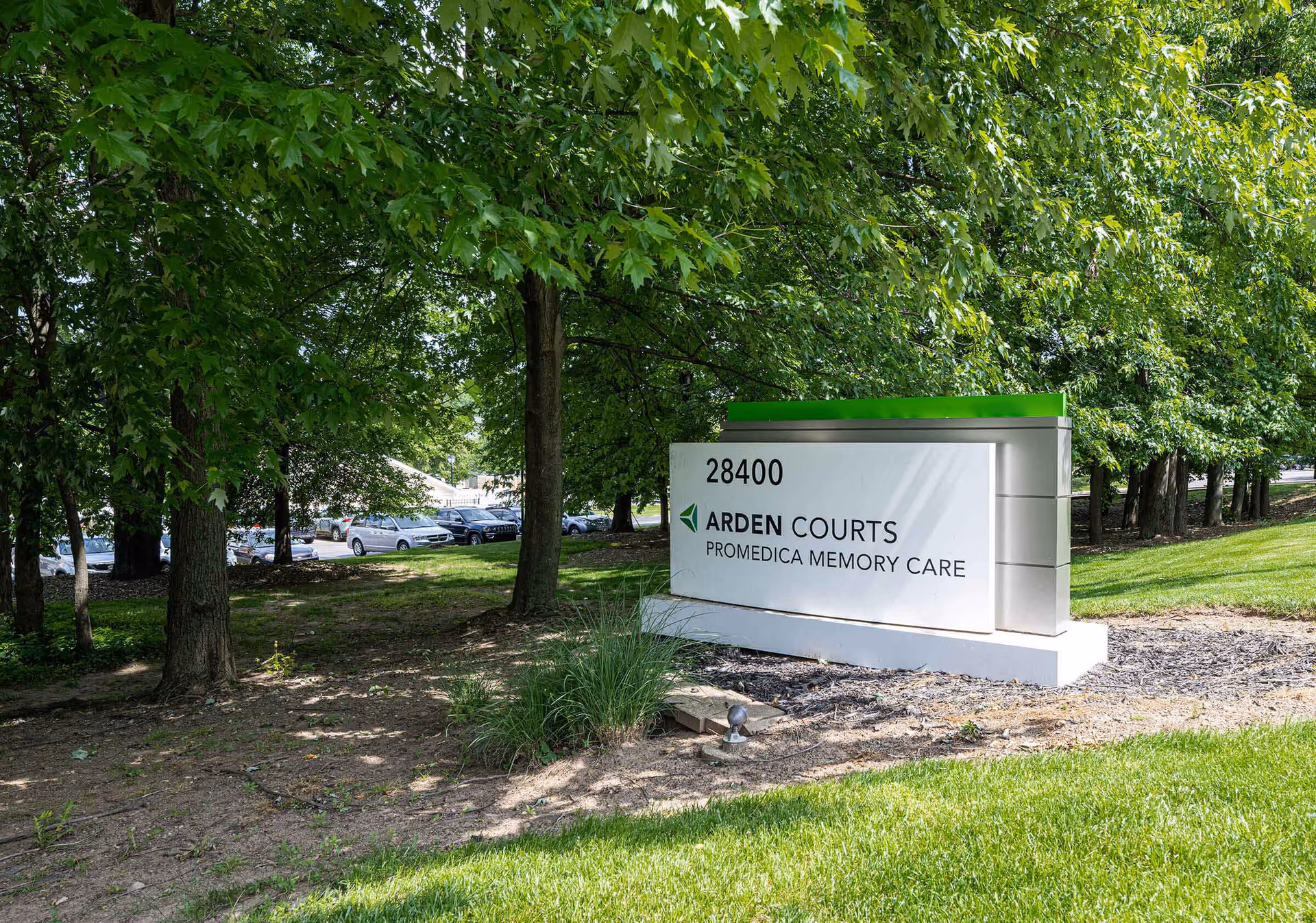 An outdoor landscaped sign reading "28400 Arden Courts ProMedica Memory Care" surrounded by trees and grass.