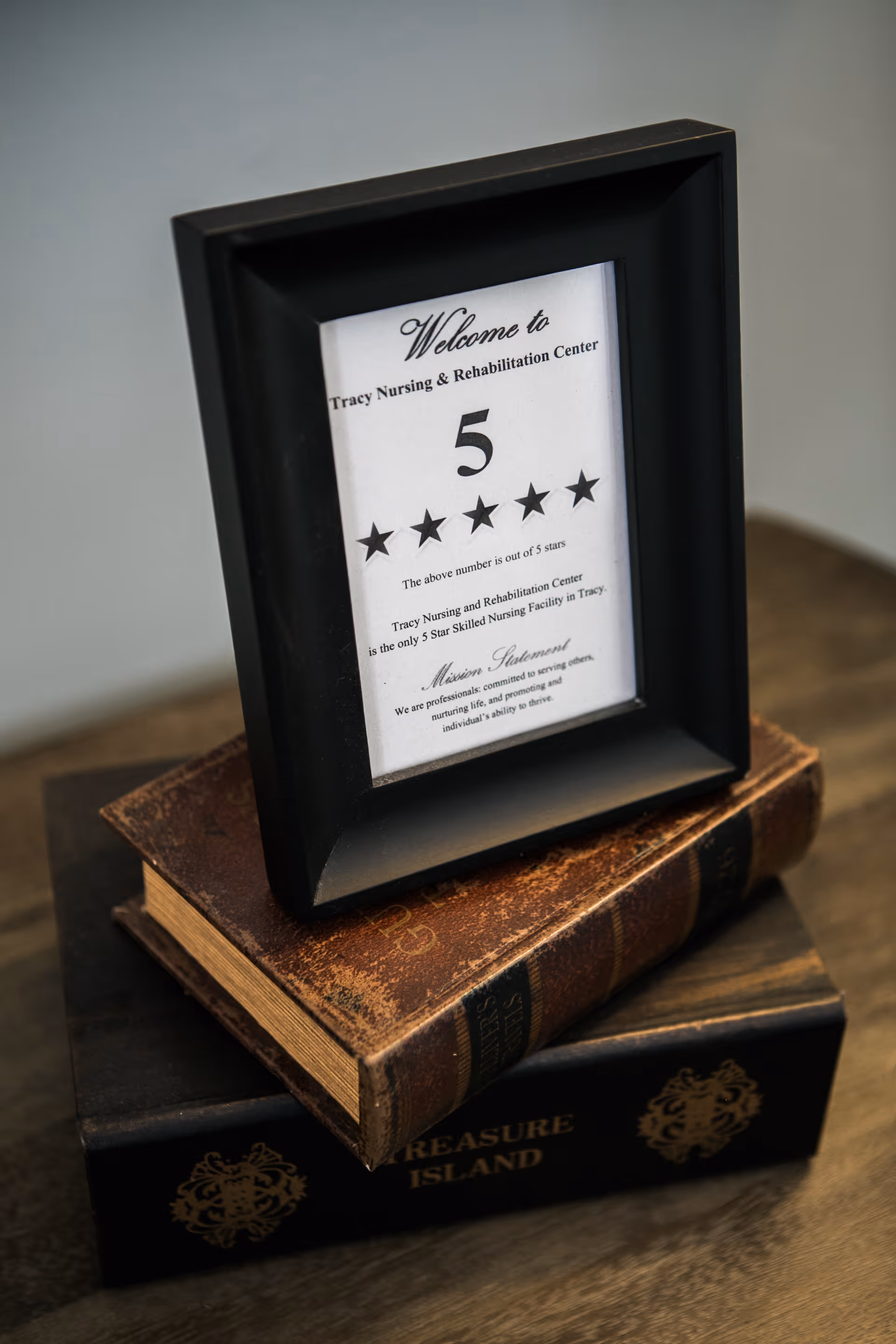 A black picture frame displaying a certificate that reads 'Welcome to Tracy Nursing & Rehabilitation Center' with a 5-star rating. The frame is placed on top of two stacked books, one titled 'Treasure Island'.