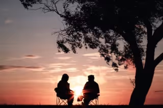 Two silhouetted people sit in lawn chairs under a tree at sunset.