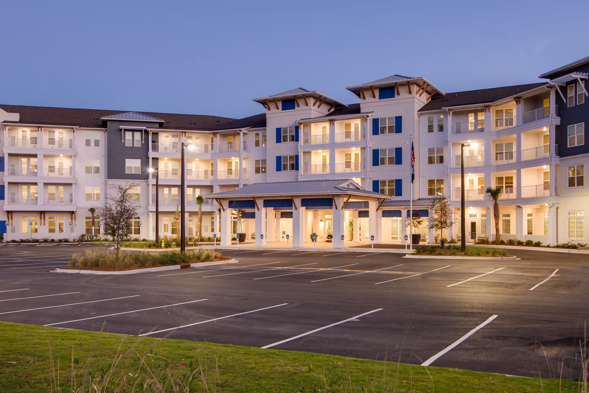 Exterior view of a large, modern senior living facility building named Portside at Grande Dunes during dusk with lights on inside. The building has multiple floors with balconies and a covered entrance. The parking lot in front is empty and well-lit.