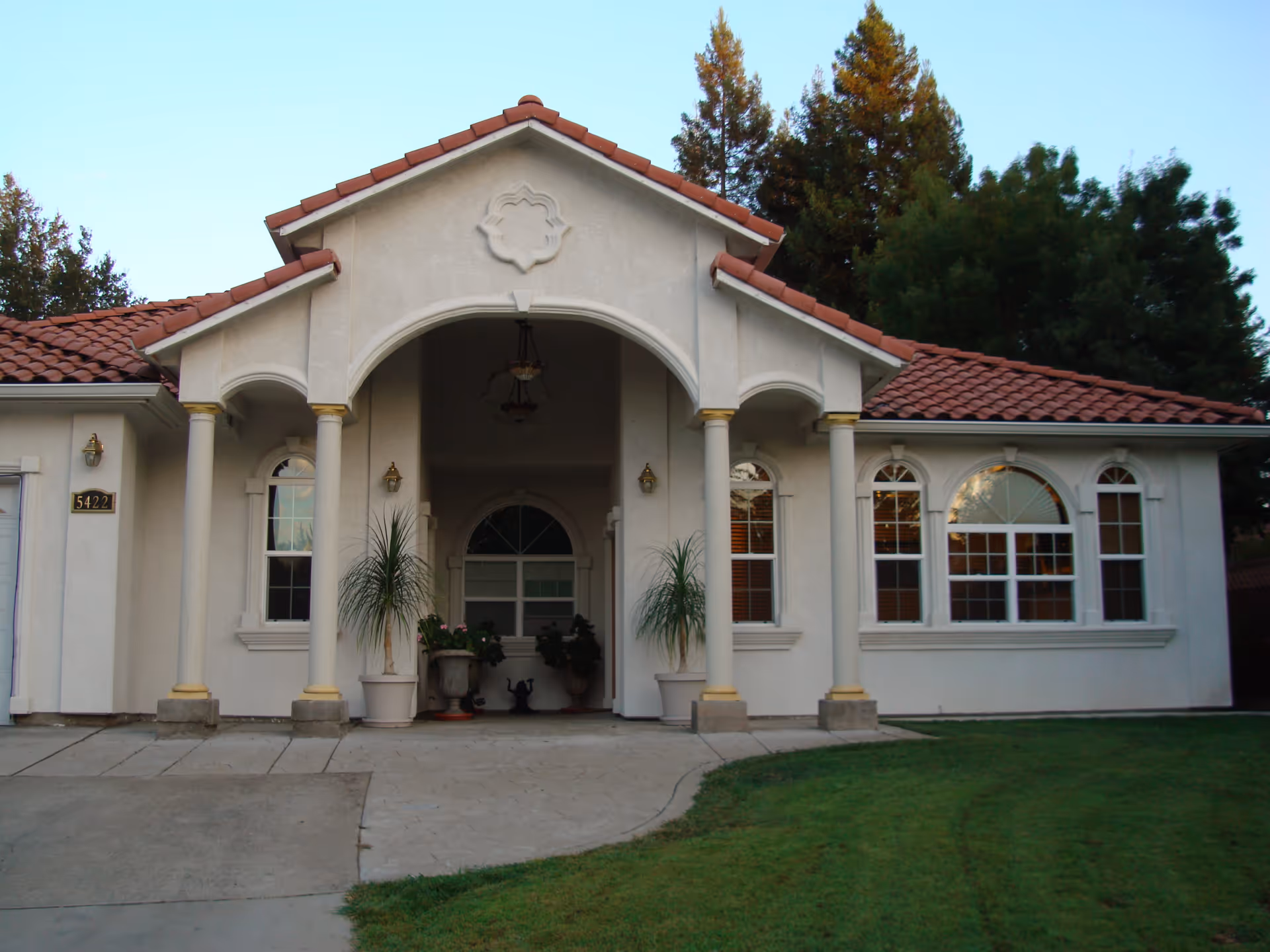 Front exterior view of a single-story building with a tiled roof, arched entrance supported by columns, several windows, and a well-maintained lawn in front.
