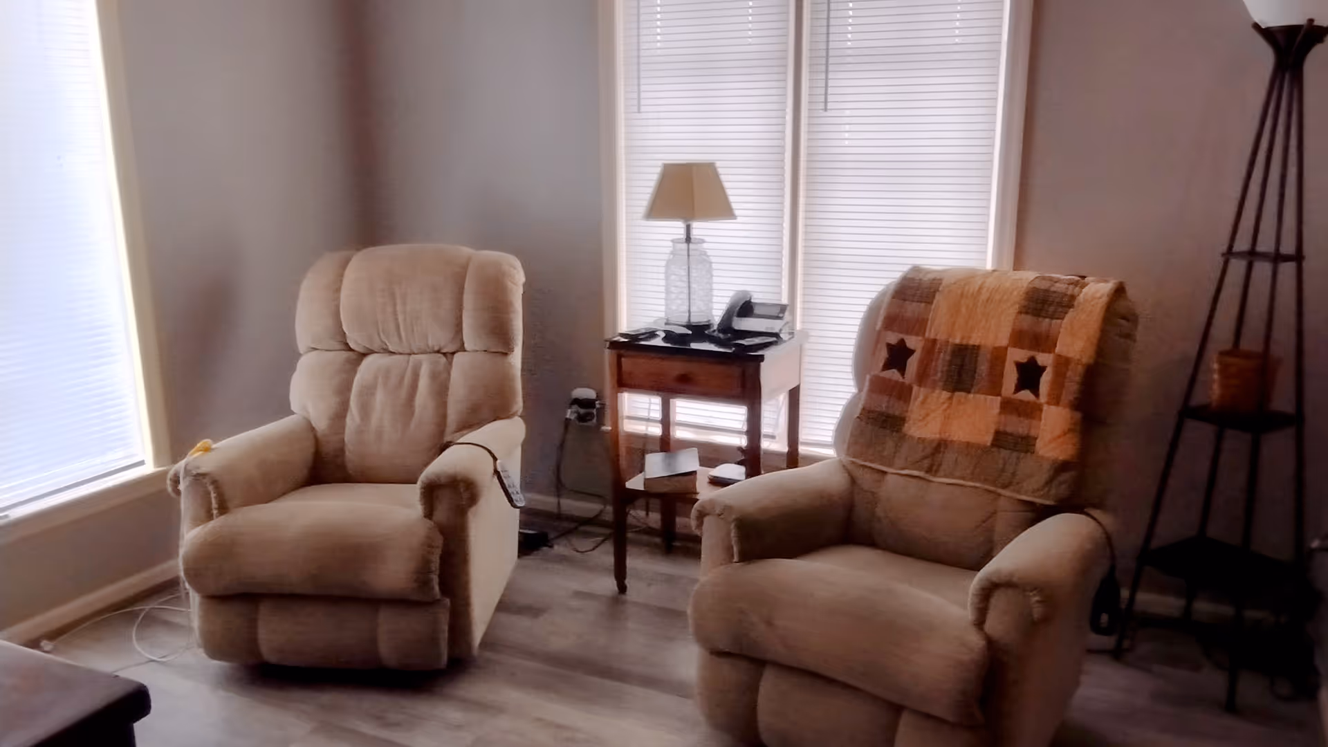 Two beige recliners sit in a cozy living room with a side table and lamp.