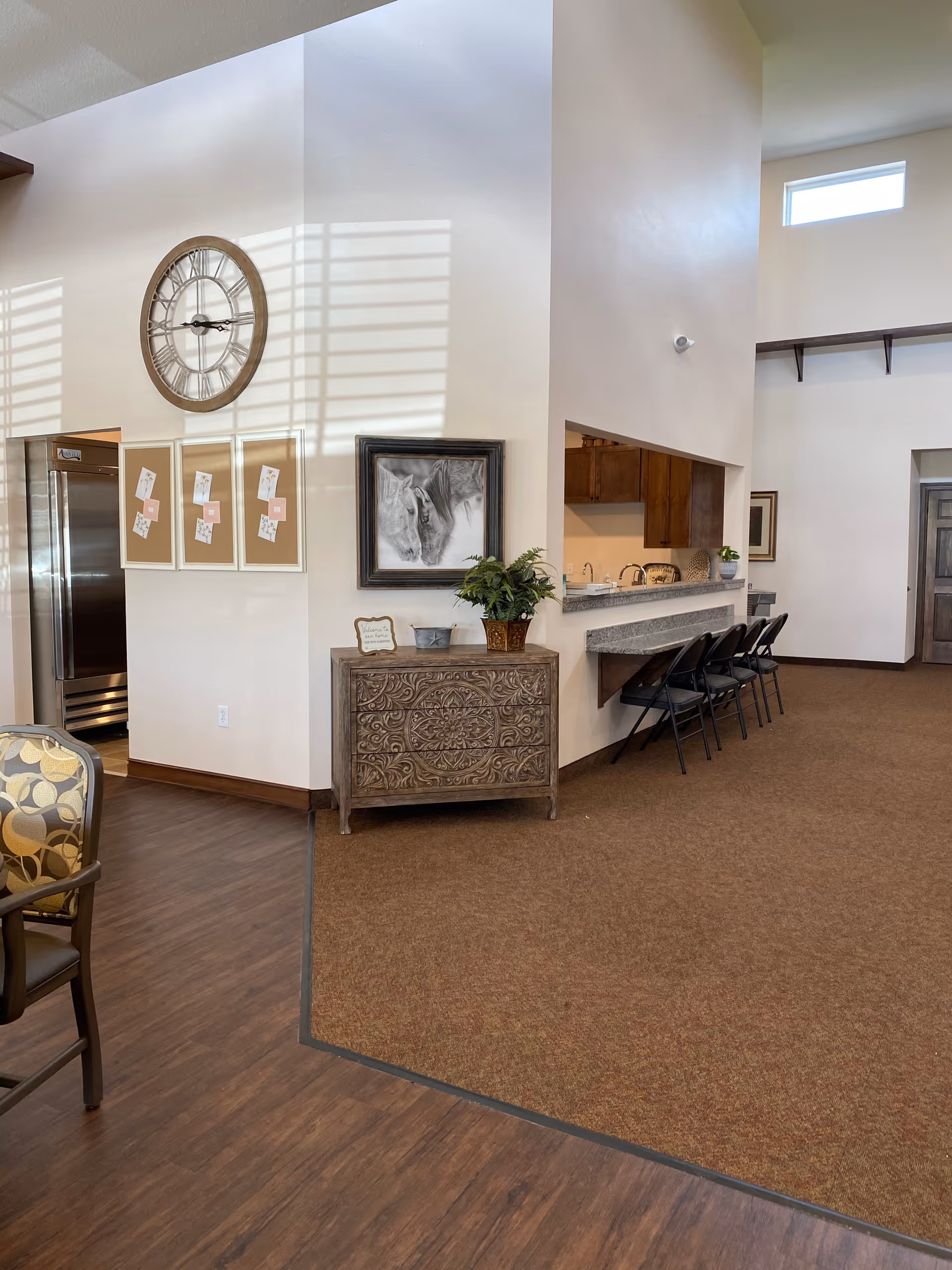 Interior view of a senior living facility showing a corner with a large wall clock, three framed bulletin boards, a decorative chest with a plant and artwork above it, and a counter with four black chairs. The floor transitions from wood to carpet, and there is a doorway leading to another room.