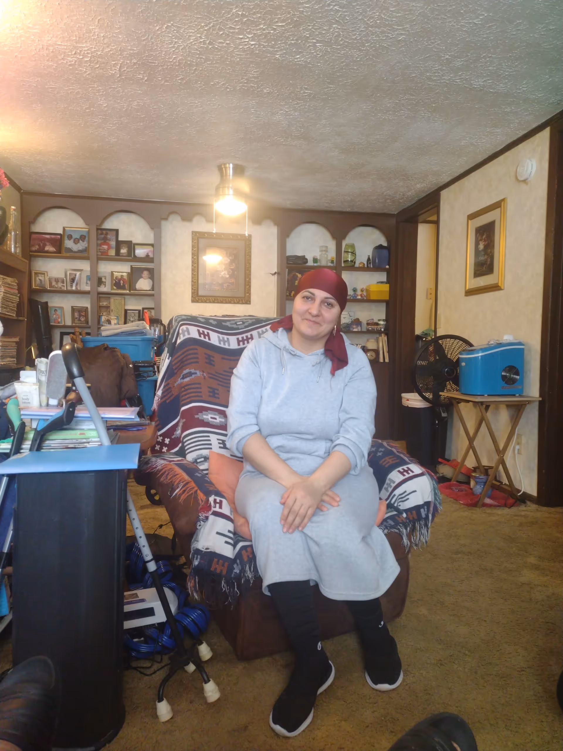 A resident sits on a patterned armchair in a cluttered living room.