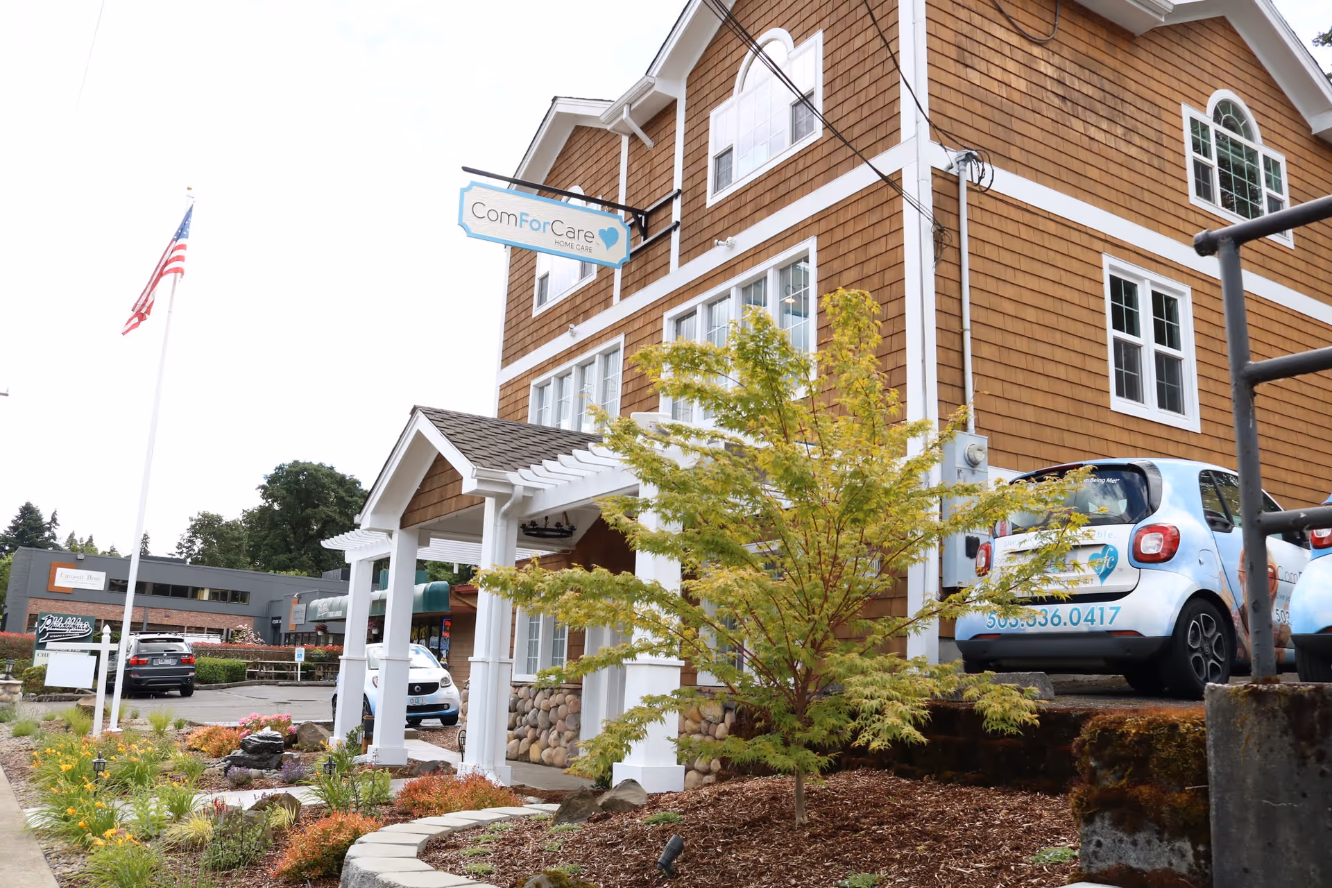 Two-story brown building with a hanging ComForCare sign, parked cars, and landscaped front yard.