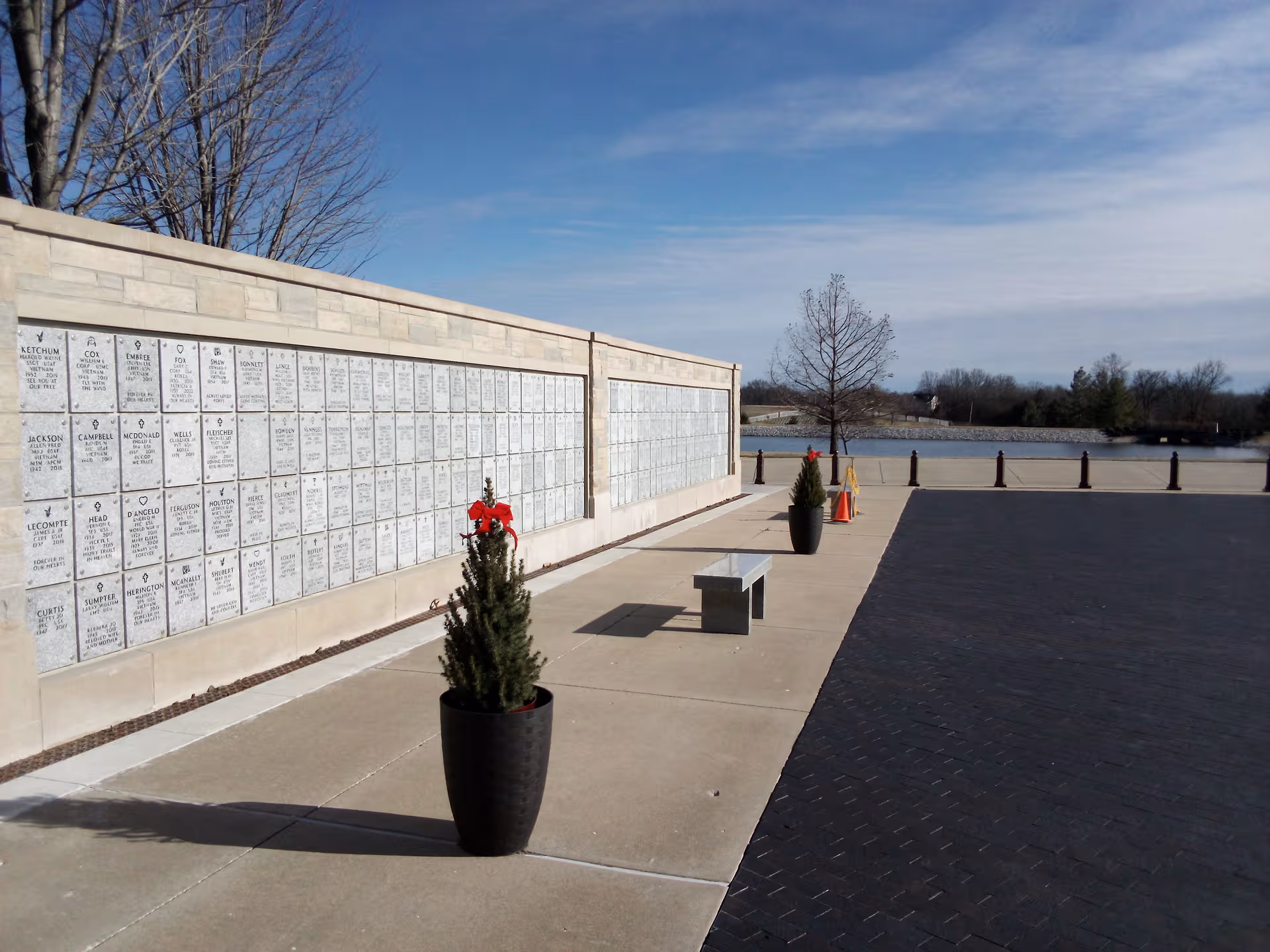 Outdoor walkway beside a memorial plaque wall with benches and potted evergreens.