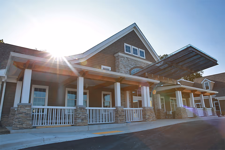 Exterior view of a senior living facility building with a covered entrance, stone and wood siding, white railings, and large windows, with sunlight shining from behind the roof.