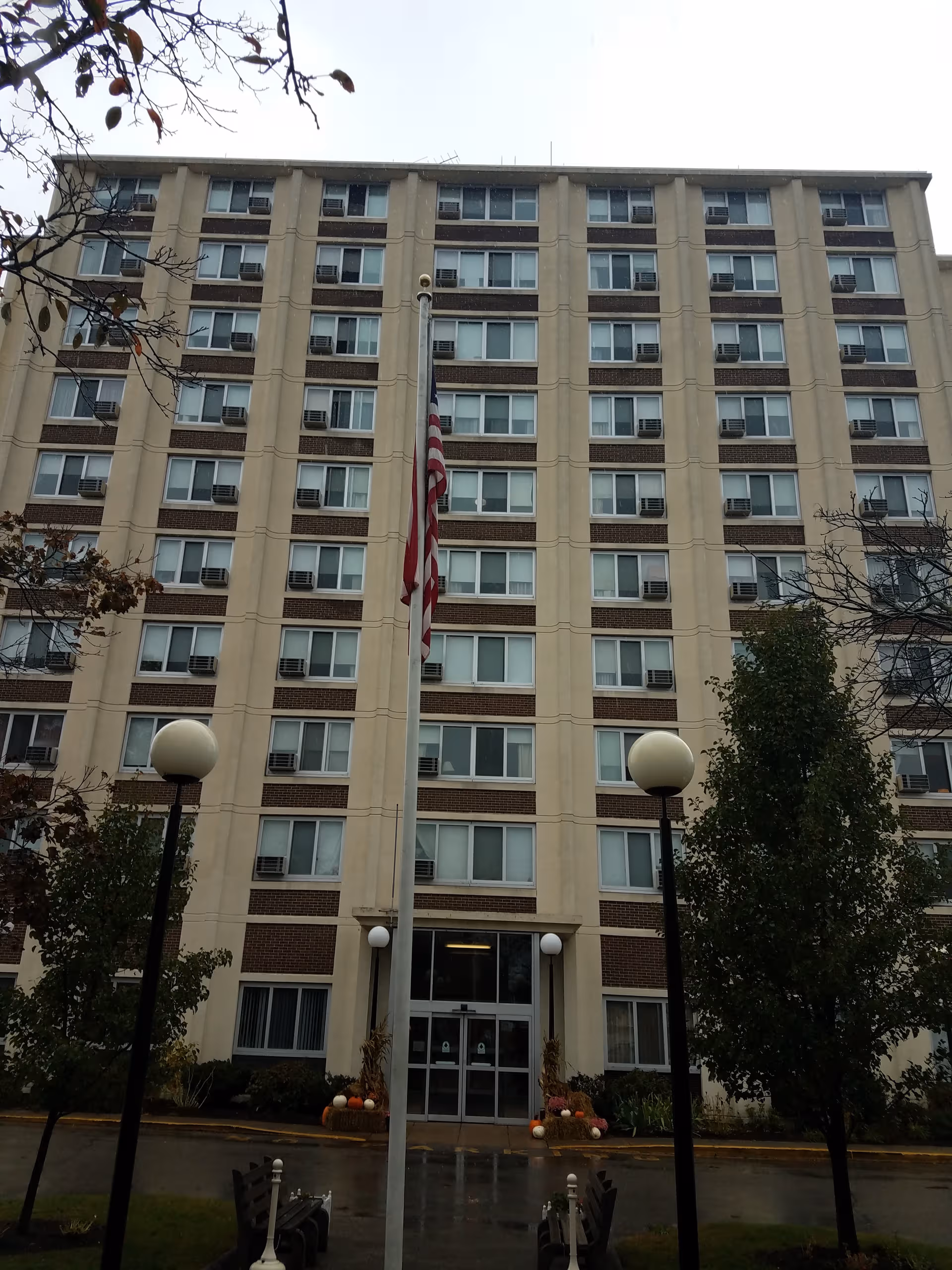 Five-story beige senior living building with flagpole, entrance, and landscaping in front.