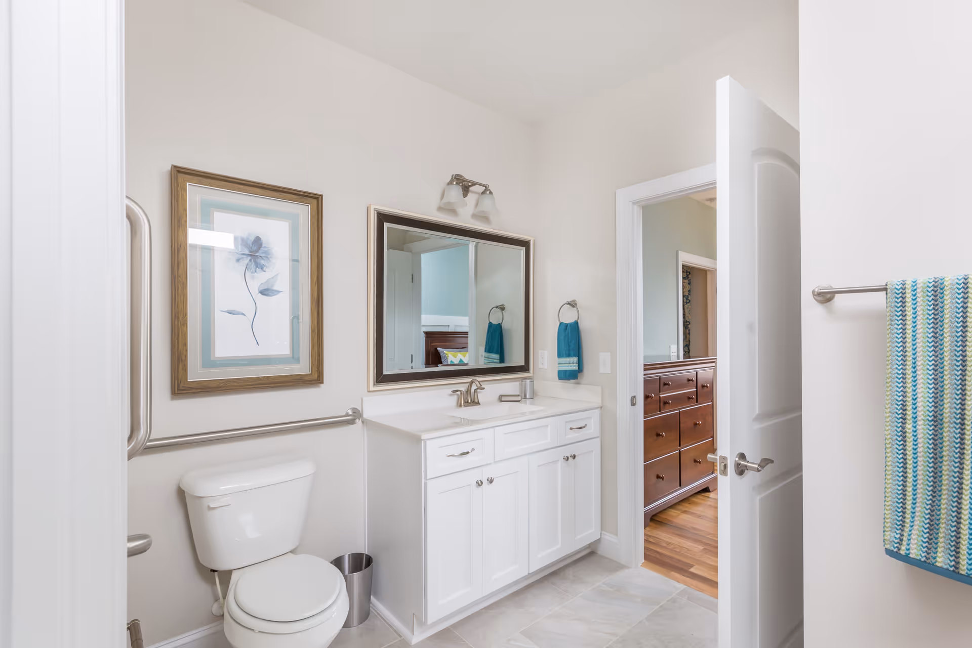 A clean and bright bathroom featuring a white toilet, a white vanity with a sink, a large mirror above the vanity, and a towel ring with a blue hand towel. There is a framed floral artwork on the wall above the toilet and a striped towel hanging on a towel bar near the door. The bathroom has light-colored walls and tile flooring. The door is open, showing a glimpse of a bedroom with wooden furniture and hardwood floors.