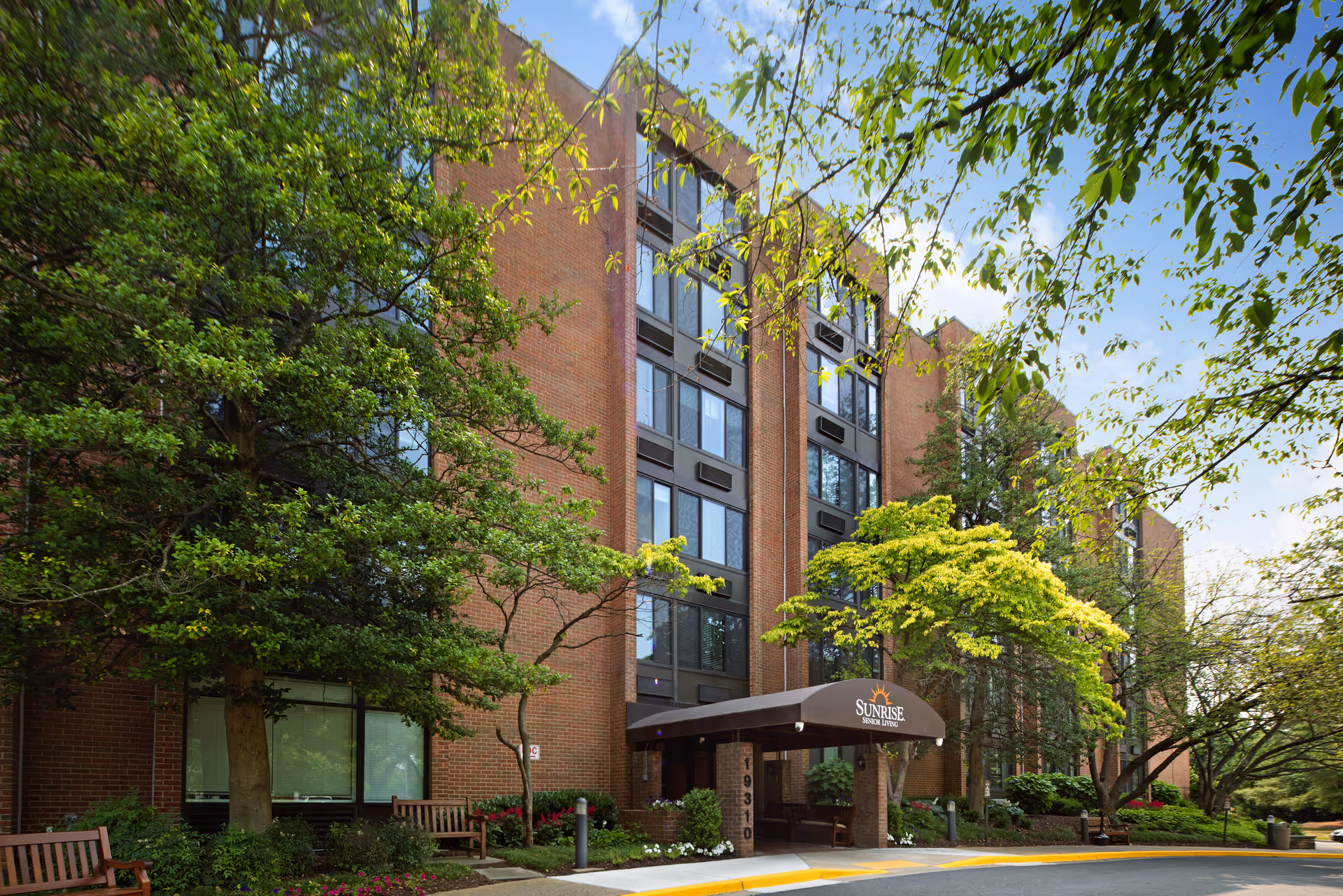 Brick senior living building with a covered entrance, trees, and a sidewalk plaza.
