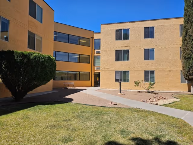Exterior view of a beige multi-story senior living building with a paved courtyard, walkway, and benches.
