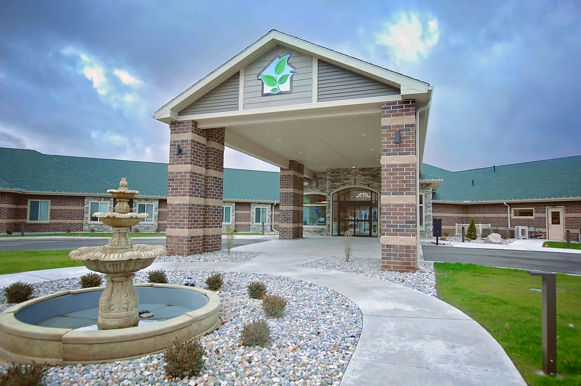 Entrance of a senior living facility with a covered driveway supported by brick pillars, a decorative fountain surrounded by rocks and small bushes, and a building with green roofs under a cloudy sky.