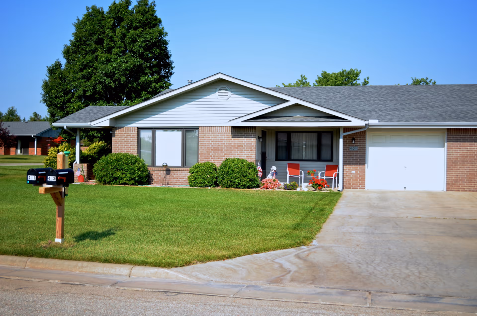 Single-story brick house with a gray roof, a small front porch with two red chairs and potted plants, a driveway leading to a white garage door, green lawn, bushes, and two black mailboxes on a wooden post near the street.