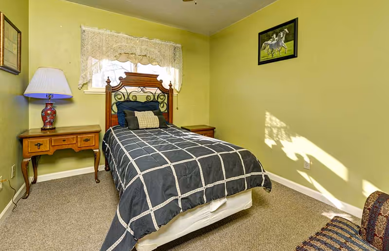 A small bedroom featuring a single bed, two nightstands, a lamp, and lace-curtained window.