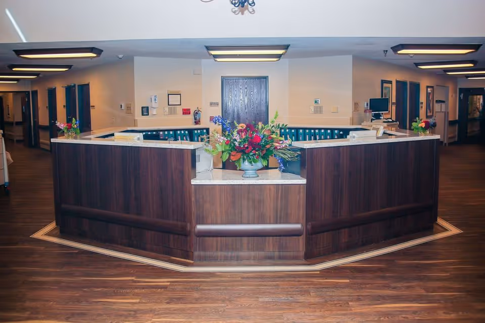 Reception desk area inside a nursing and rehabilitation center with a large wooden counter, floral arrangements on the counter, computer monitors, and hallways leading to other rooms in the background.