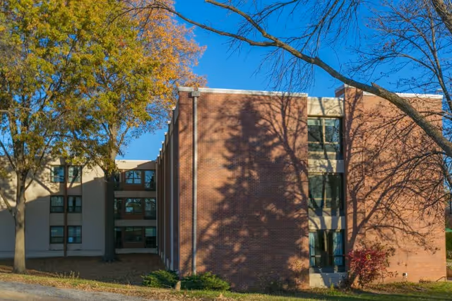 Exterior view of a multi-story brick and stucco building with trees casting shadows under a clear blue sky.