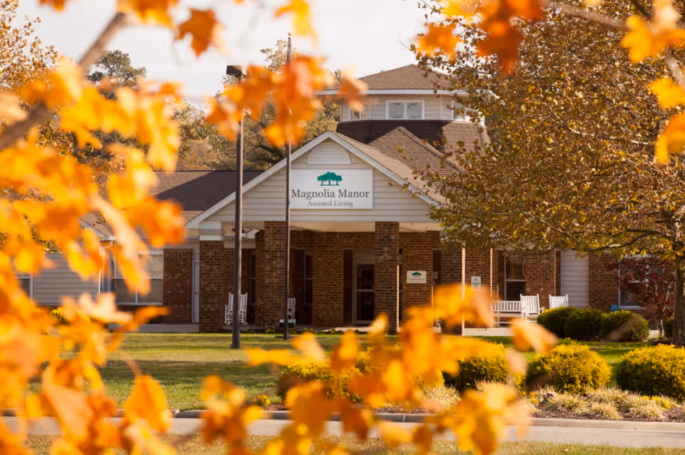 Front exterior view of Magnolia Manor Assisted Living building with autumn foliage in the foreground and a well-maintained lawn and bushes.