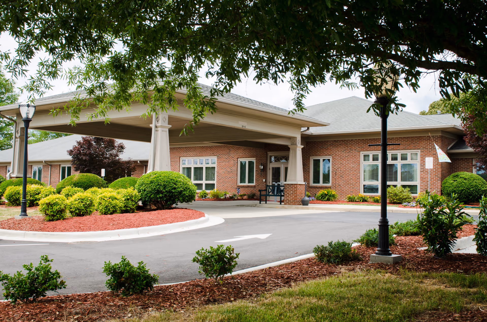 Covered porte-cochère and entrance of a brick senior living building surrounded by landscaped shrubs and trees.