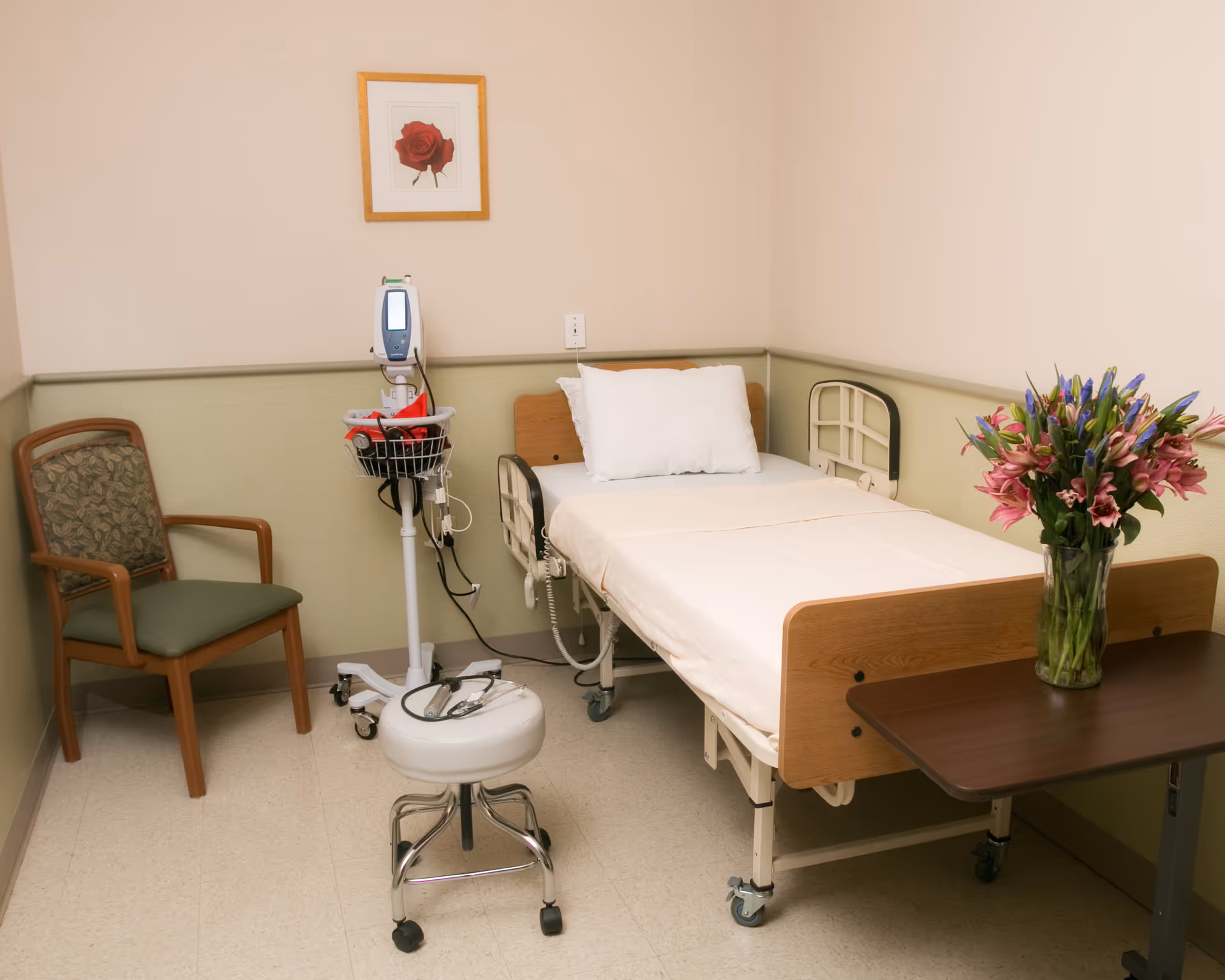 Hospital-style patient room with bed, monitor, chair, and vase of flowers.