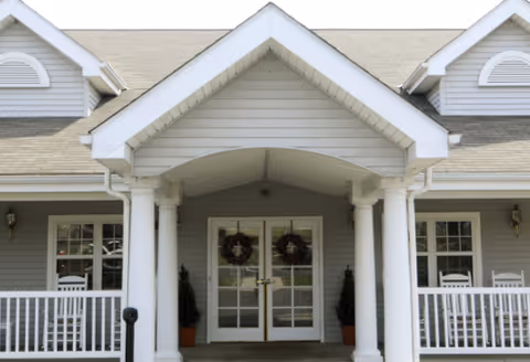 Front entrance of a building with a covered porch supported by white columns, double glass doors decorated with wreaths, and a gray exterior with white trim. There are rocking chairs on the porch on either side of the entrance.