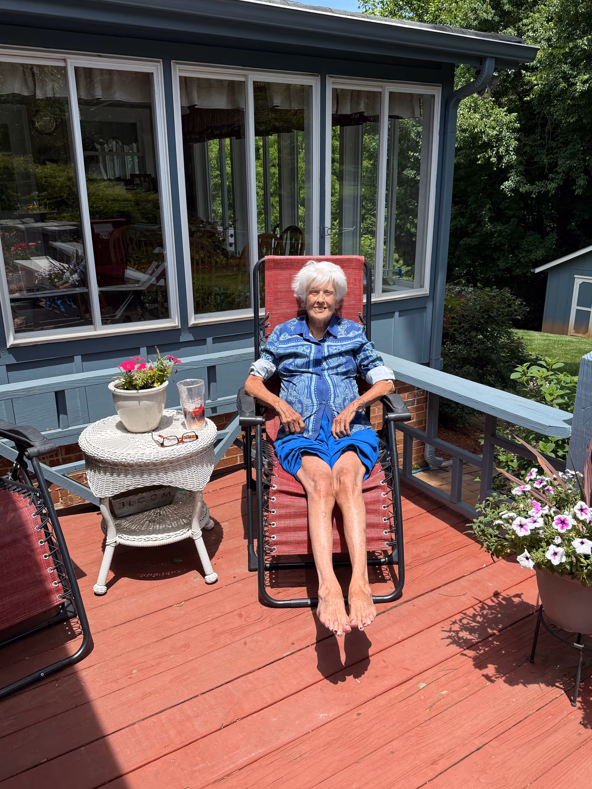 Elderly woman sits in a red lounge chair on a sunny outdoor deck beside potted flowers.