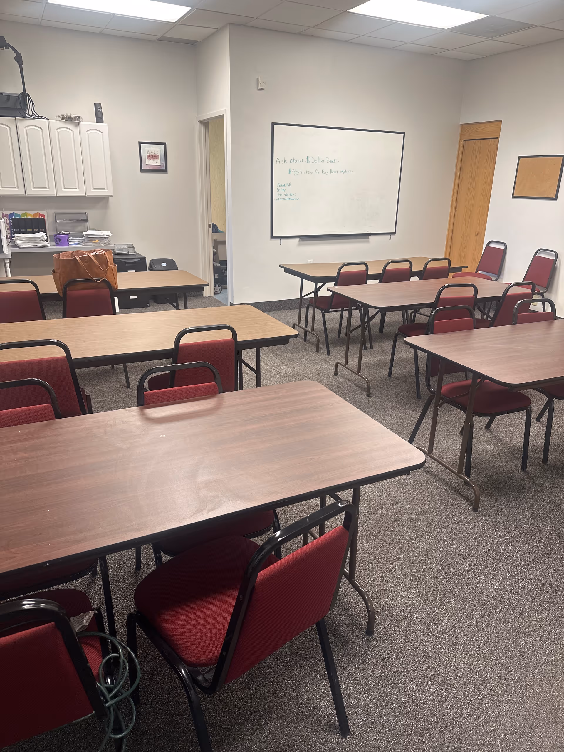 Interior classroom-style meeting room with tables, red chairs, a whiteboard, and cabinets.