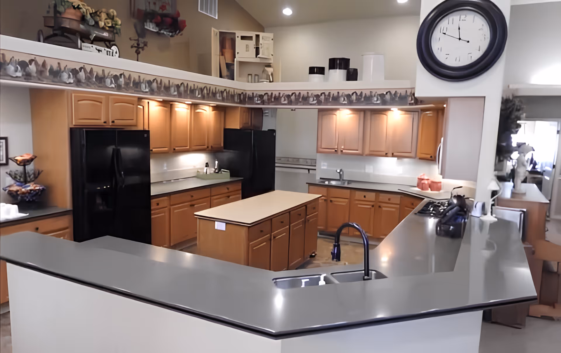 A spacious kitchen with wooden cabinets, black appliances including two refrigerators, a central island, and a large gray countertop with a sink and faucet. The kitchen is well-lit with under-cabinet lighting and recessed ceiling lights. A large black wall clock is mounted on a white pillar, and decorative items are placed on the counters and shelves.