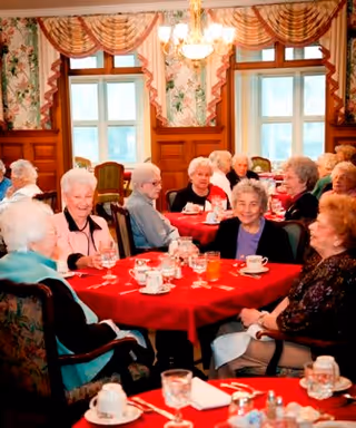 A group of elderly residents seated around tables with red tablecloths in a decorated dining room, enjoying drinks and conversation.